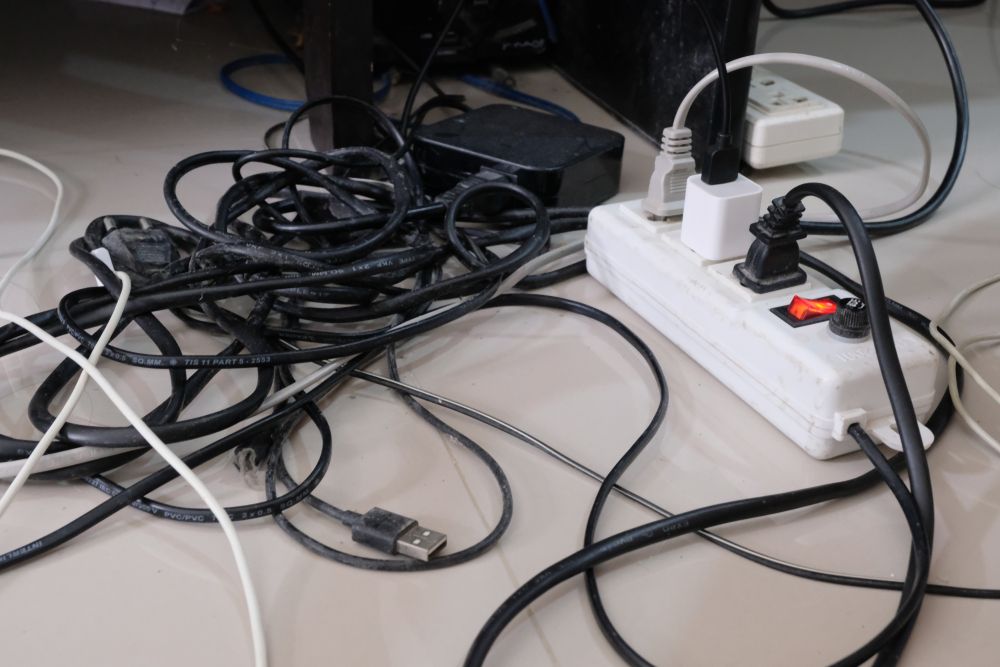 multiple electrical plugs and wires tangled under a desk and plugged into one power bar