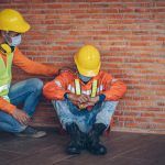 2 workers sitting by a brick wall with one of them comforting the other who is exhausted