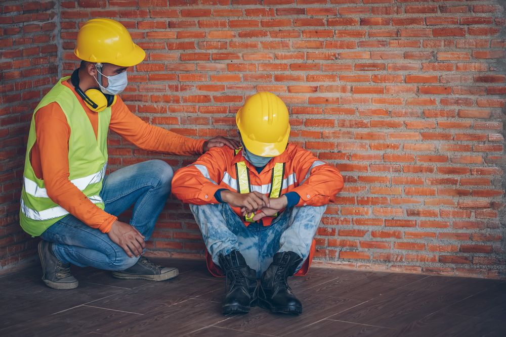 2 workers sitting by a brick wall with one of them comforting the other who is exhausted