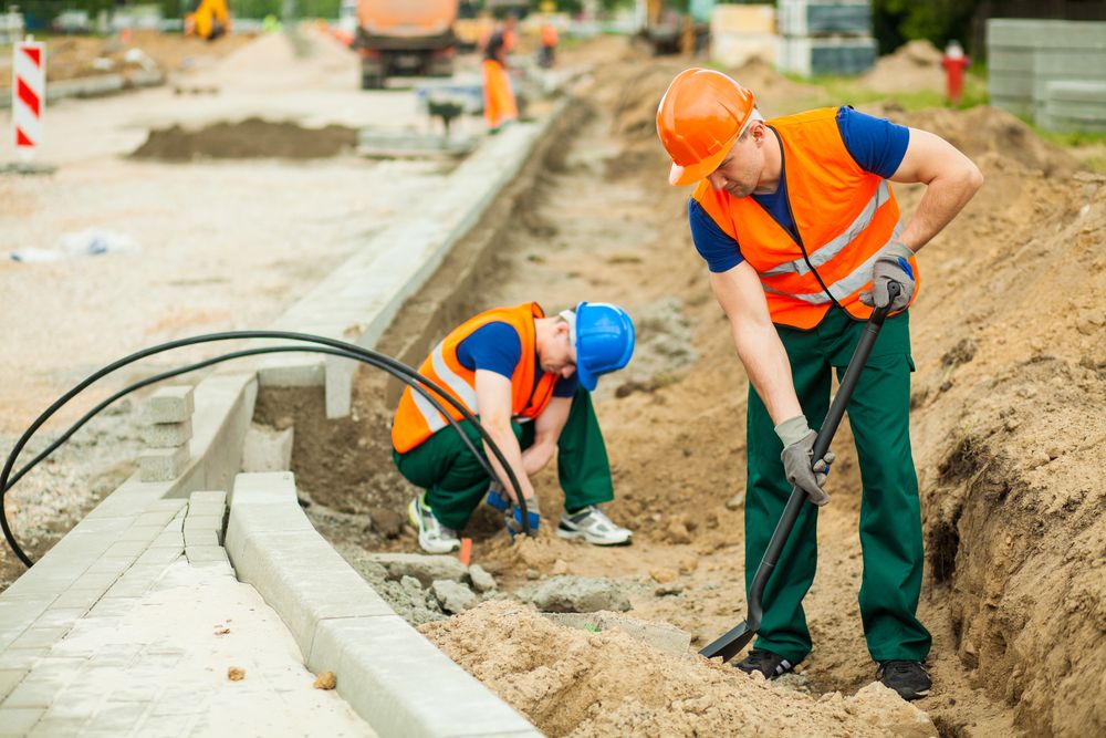 two construction workers on a site, one is using a shovel and the other is laying wires into the ground.